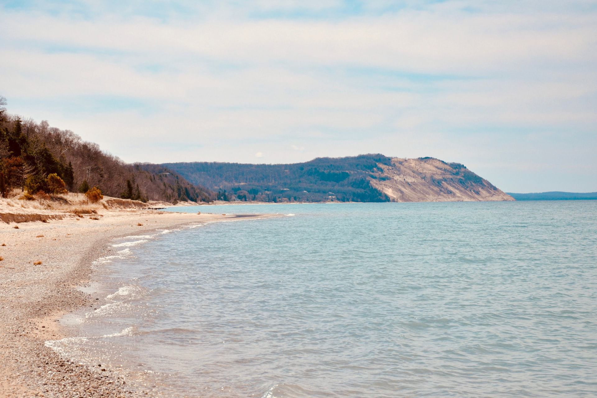 A wide view of the Lake Michigan shoreline curving toward a tree-covered bluff, with gentle ripples along a sandy and pebbled beach under a soft sky