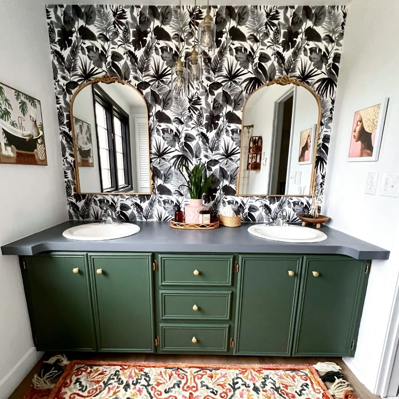 A bathroom designed by the Openwater team with black-and-white tropical wallpaper, a sage-green double vanity, and arched brass mirrors