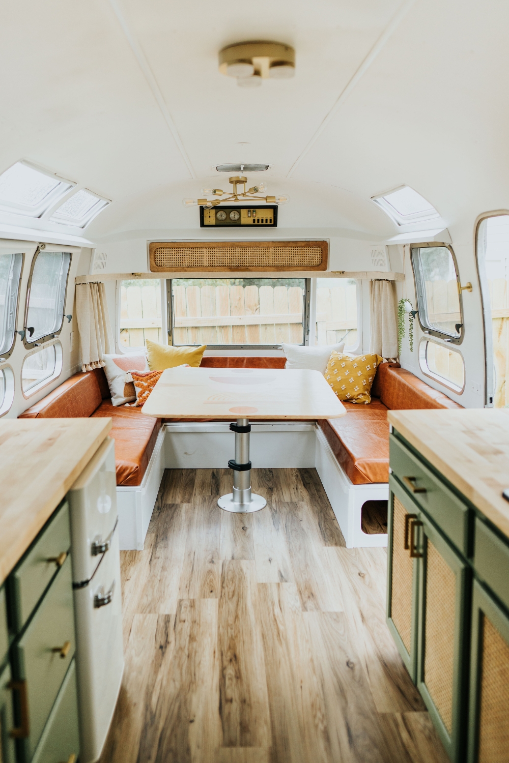 A renovated Airstream interior with a cognac leather dinette booth, sage-green cabinets, warm wood floors, and brass light fixtures