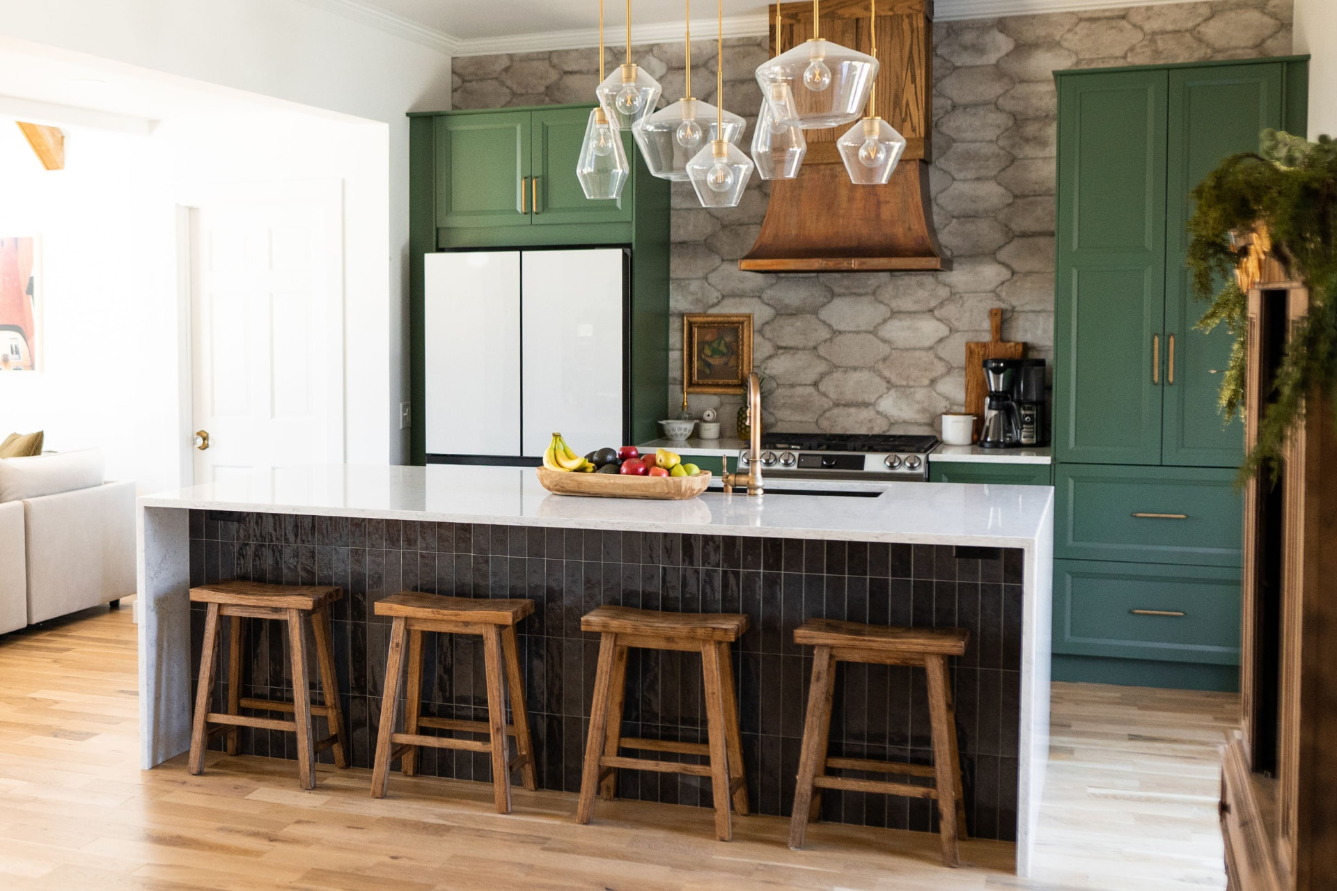 A kitchen renovated by the Openwater team with forest-green cabinetry, a stone tile backsplash, glass pendant lights, and a white waterfall island with wooden stools