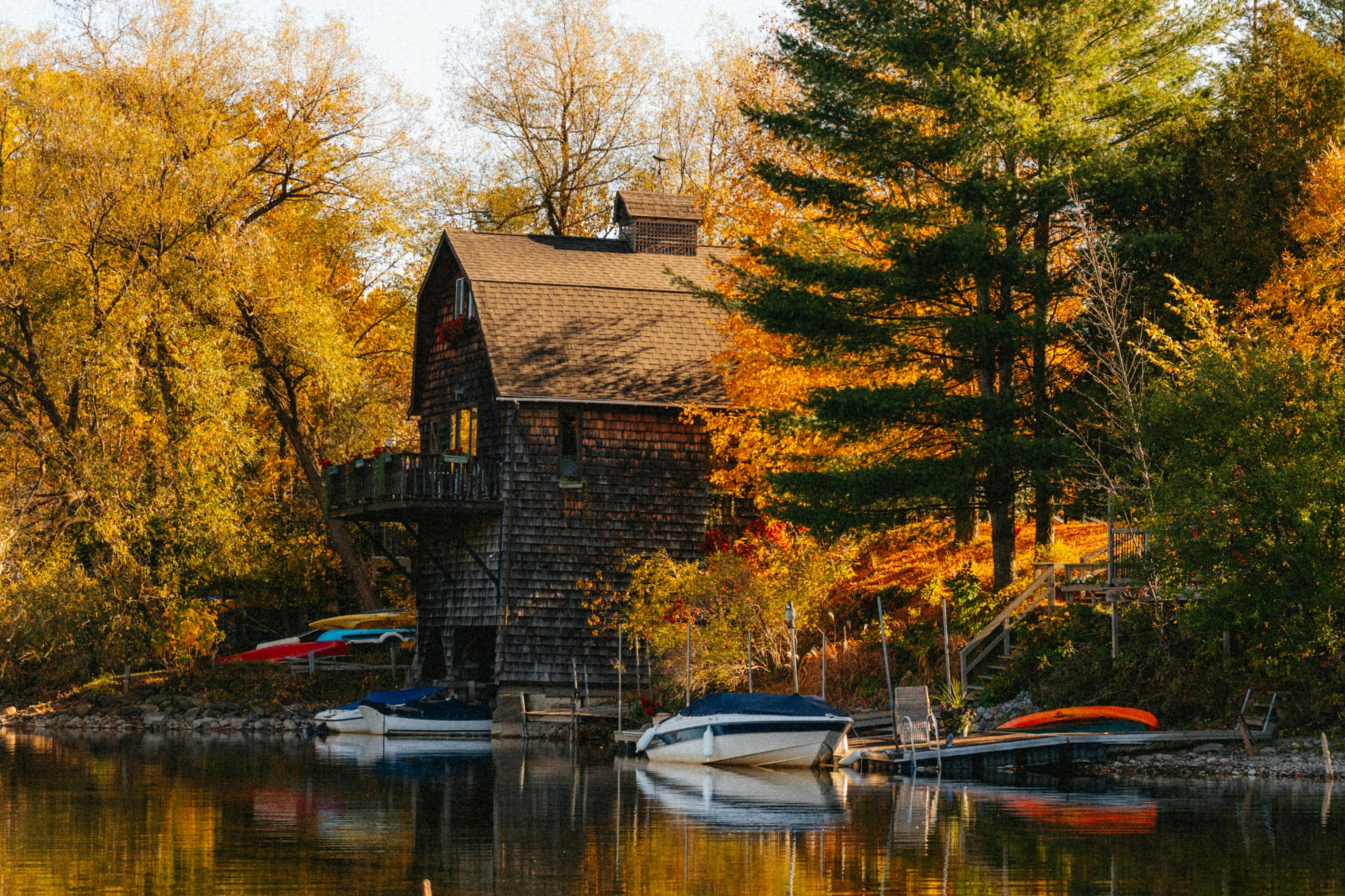 A weathered wooden boathouse on a calm Lake Michigan inlet, framed by vivid autumn foliage with small boats docked at the water's edge