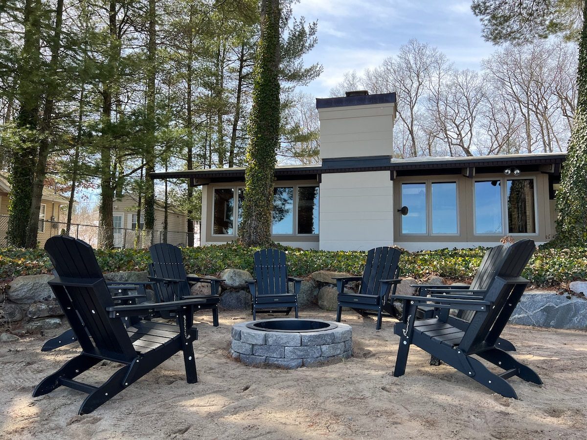 Black Adirondack chairs around a stone fire pit on the sand beside the white MCM Lake House in Twin Lake