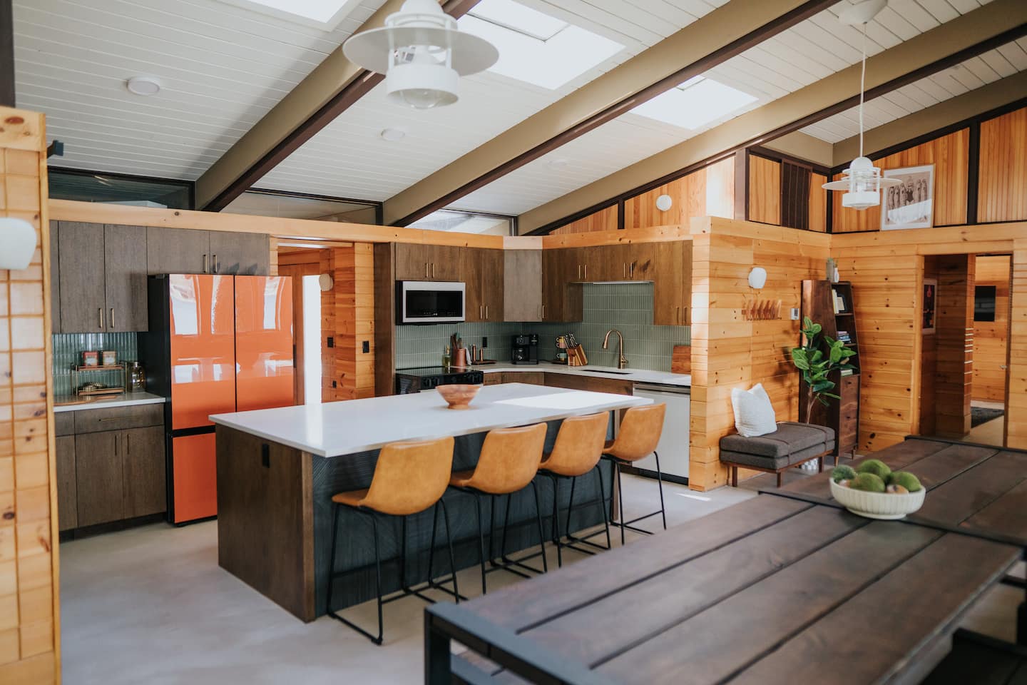 The mid-century modern kitchen at the MCM Lake House, an Open Water co-hosted home, with warm wood paneling, leather bar stools, and a bold orange refrigerator